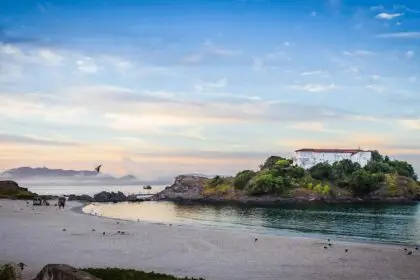 O que fazer em cabo frio: Praia do Forte em Cabo Frio ao entardecer com Forte São Mateus na ilha, águas calmas, areia branca, céu dourado e pássaros voando