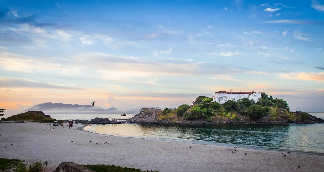 O que fazer em cabo frio: Praia do Forte em Cabo Frio ao entardecer com Forte São Mateus na ilha, águas calmas, areia branca, céu dourado e pássaros voando