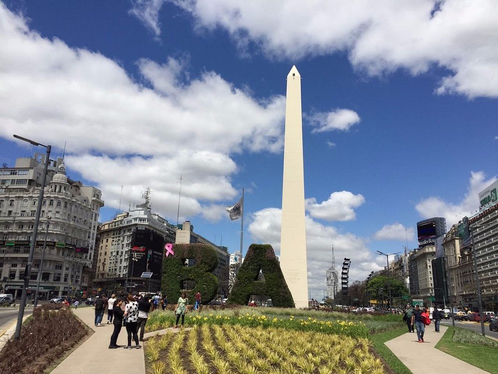 Obelisco de Buenos Aires em dia ensolarado, com o letreiro "BA" coberto por plantas e pessoas caminhando ao redor.