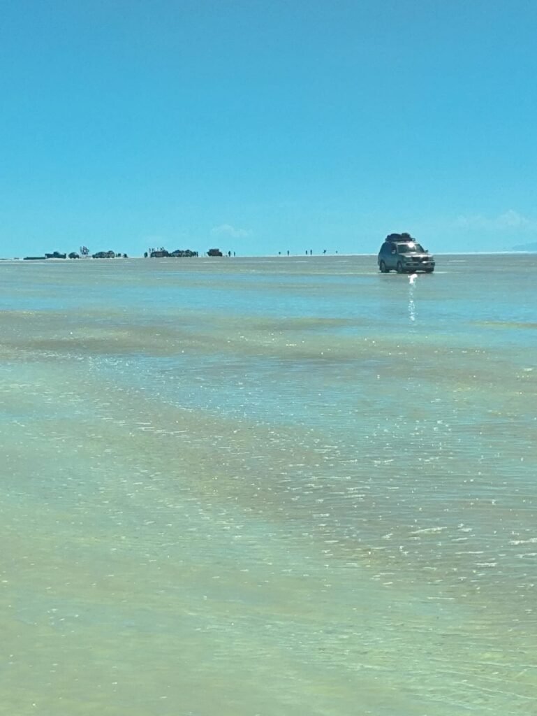 Panorâmica do Salar de Uyuni na época chuvosa com veículo 4x4 sobre espelho d'água refletindo céu azul e nuvens brancas